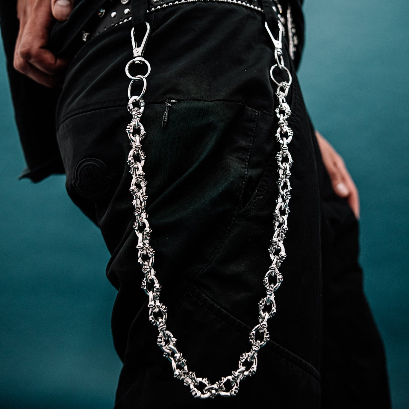 Person wearing a black belt with studs and a silver chain, against a dark background