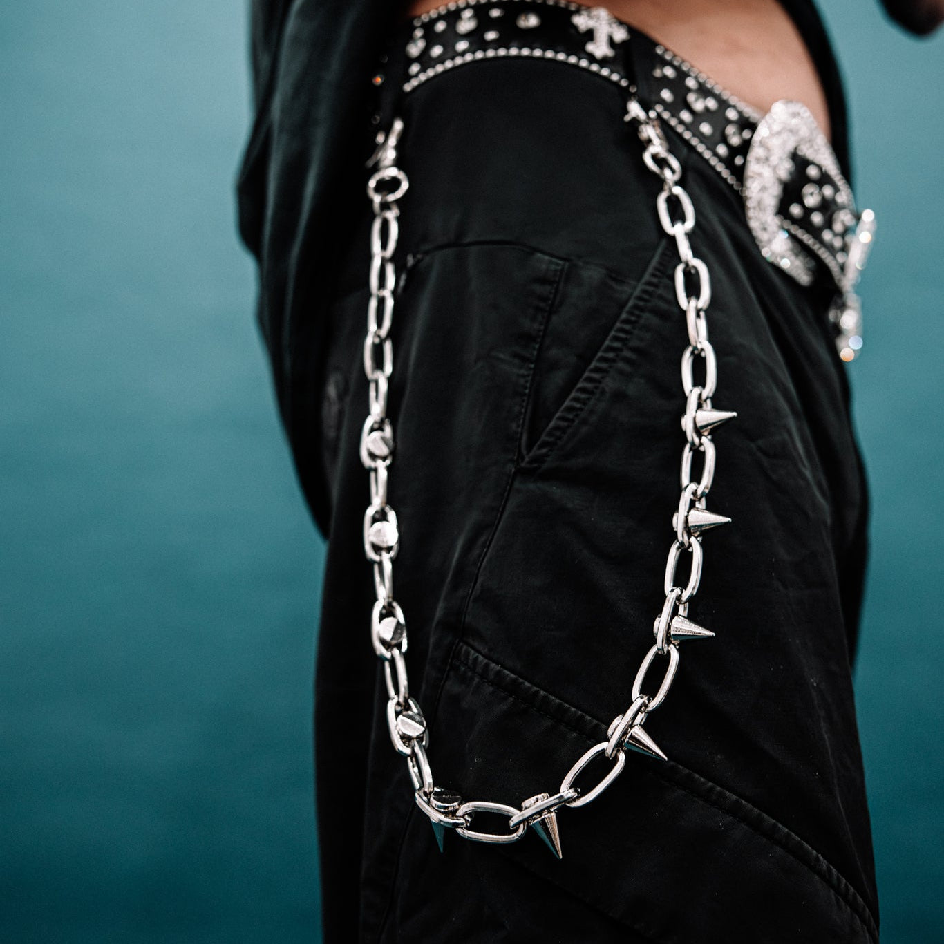 Person wearing a black outfit with a chain necklace and studded details against a teal background